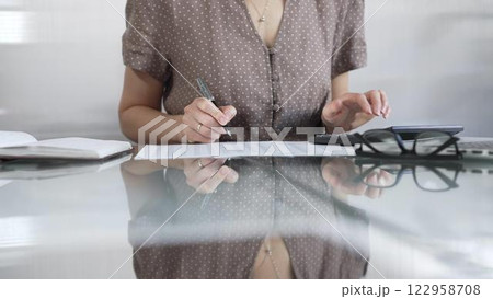 Businesswoman accountant, wearing brawn dotted blouse, is using a calculator to crunch numbers and jotting down meticulous notes, at sleek glass desk in a contemporary office. Taxes, audit in business Businesswoman accountant, wearing brawn dotted blouse, is using a calculator to crunch numbers and jotting down meticulous notes, at sleek glass desk in a contemporary office. Taxes, audit in business 122958708