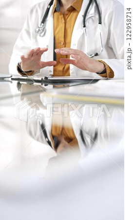 Unknown male doctor wearing a stethoscope, yellow shirt under a lab coat is gesturing with hands overs a glass desk with a laptop in clinic office. Medicine concept 122958714
