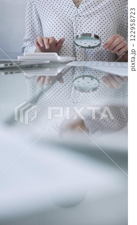 Businesswoman in white dotted blouse is carefully reviewing financial reports with magnifying glass and calculator, conducting audit and analyzing data, vertical view Taxes, audit in business Businesswoman in white dotted blouse is carefully reviewing financial reports with magnifying glass and calculator, conducting audit and analyzing data, vertical view Taxes, audit in business 122958723