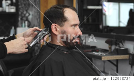 Skilled barber trimming male client's hair using electric clipper, creating contemporary hairstyle in modern urban barbershop setting with precise grooming technique 122958724
