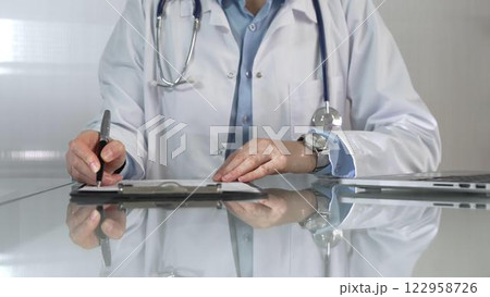 Woman doctor, wearing a blue blouse under a white medical coat, is using a clipboard and taking notes on glass desk in a medical office, close-up. Medicine 122958726