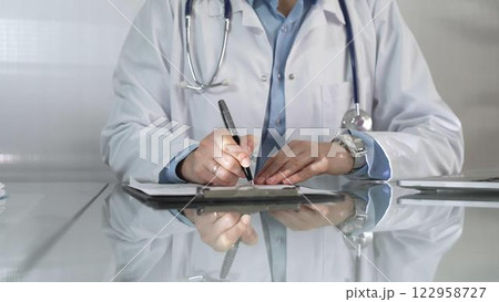 Woman doctor, wearing a blue blouse under a white medical coat, is using a clipboard and taking notes on glass desk in a medical office, close-up. Medicine 122958727