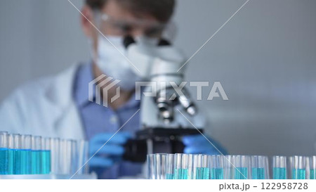 Laboratory tubes are in focus, closeup view. Man scientist researcher with a blue protective gloves is using a microscope at the background. Medicine and science concept 122958728