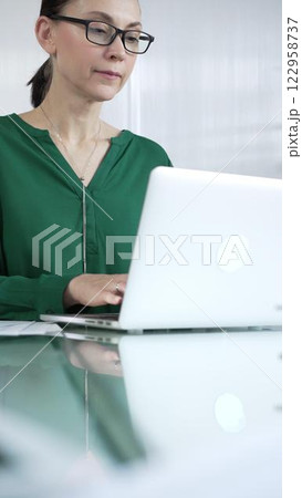 Businesswoman, wearing green blouse, is typing on laptop keyboard, preparing taxes and audit reports, with documents on glass office desk, vertical portrait view. Taxes, audit in business Businesswoman, wearing green blouse, is typing on laptop keyboard, preparing taxes and audit reports, with documents on glass office desk, vertical portrait view. Taxes, audit in business 122958737