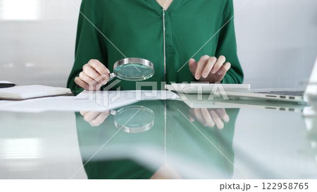 Businesswoman in green blouse carefully reviewing financial reports with magnifying glass and calculator, conducting audit and analyzing data in office. Taxes, audit in business Businesswoman in green blouse carefully reviewing financial reports with magnifying glass and calculator, conducting audit and analyzing data in office. Taxes, audit in business 122958765