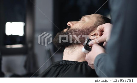 Professional barber trimming client's beard with electric razor, delivering precise styling within contemporary grooming salon setting 122958781