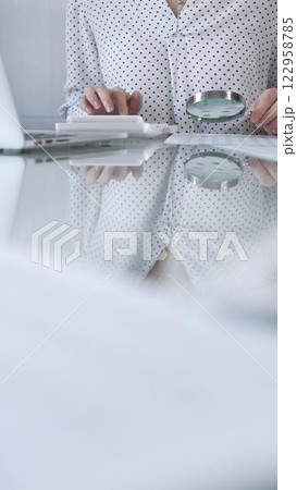 Businesswoman in white dotted blouse is carefully reviewing financial reports with magnifying glass and calculator, conducting audit and analyzing data, vertical view Taxes, audit in business 122958785