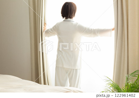 朝の寝室でカーテンを開けるパジャマ姿のミドル女性 朝の寝室でカーテンを開けるパジャマ姿のミドル女性 122961402
