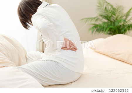 朝の寝室で腰痛に苦しむ白いパジャマ姿のミドル女性 122962533