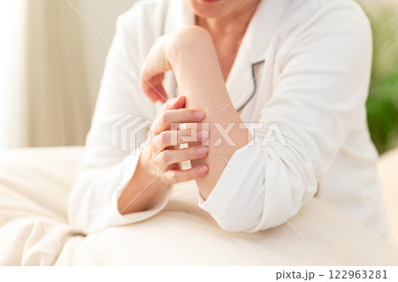 朝の寝室で腕の乾燥肌に悩む白いパジャマ姿のミドル女性 朝の寝室で腕の乾燥肌に悩む白いパジャマ姿のミドル女性 122963281