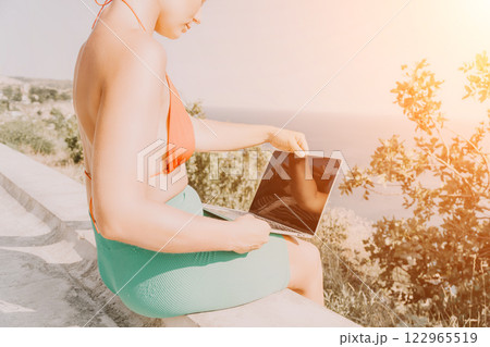 Woman Laptop Beach Working Remotely on Vacation - Beautiful young woman in a bathing suit using a laptop while sitting on a cliff overlooking the ocean. 122965519