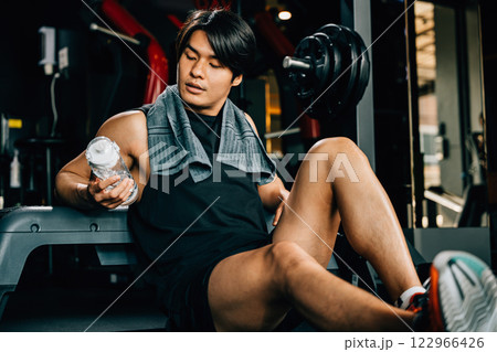 Portrait of Asian young man trained body in black sportswear sitting resting holding water or proteins on a bench after a workout in fitness gym, Weight training, healthy lifestyle concept 122966426