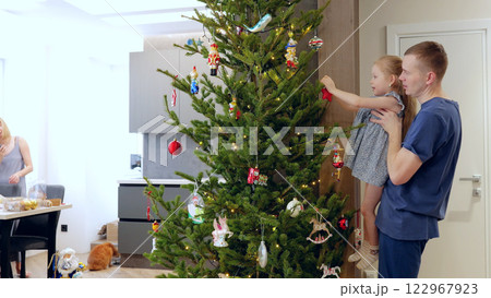 Young family is decorating a christmas tree in their home, getting ready for the holiday season. The father is holding his daughter as she hangs an ornament 122967923
