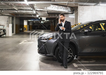 Caucasian male business professional holding money and speaking on phone in underground parking garage. Dressed in formal suit, exhibiting confidence. 122969791