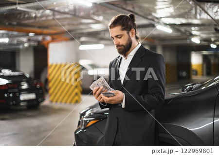 Young Caucasian man in suit counts cash in dimly lit underground parking lot next to car. 122969801