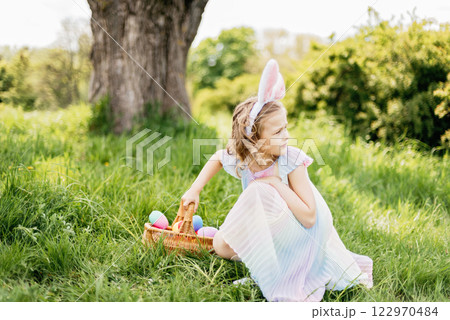 Easter egg hunt. Girl child Wearing Bunny Ears Running To Pick Up Egg In Garden. Easter tradition. Baby with basket full of colorful eggs. Bunny in basket Easter egg hunt. Girl child Wearing Bunny Ears Running To Pick Up Egg In Garden. Easter tradition. Baby with basket full of colorful eggs. Bunny in basket 122970484
