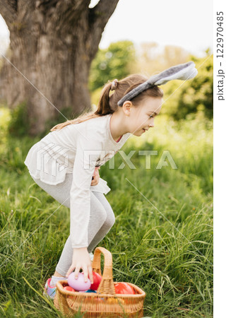 Easter egg hunt. Girl child Wearing Bunny Ears Running To Pick Up Egg In Garden. Easter tradition. Baby with basket full of colorful eggs. Bunny in basket 122970485
