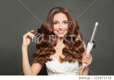 Attractive brown haired woman with shiny long curly hairstyle showing curling iron. Attractive brown haired woman with shiny long curly hairstyle showing curling iron. 122971518