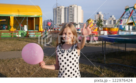 A cheerful girl with a bright smile waves at the camera, holding a colorful pink balloon at a vibrant fair 122976424