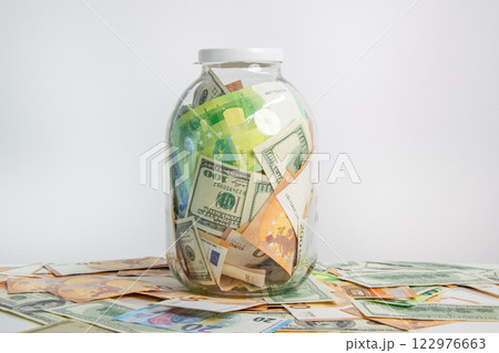 A glass jar filled with various US dollar and euro banknotes sits on a white surface, surrounded by scattered currency bills 122976663
