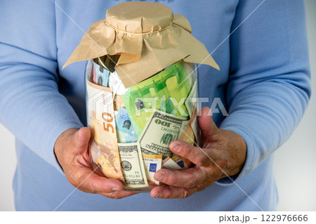 A person is holding a glass jar filled with a variety of euro and dollar bills. The jar is wrapped in brown paper A person is holding a glass jar filled with a variety of euro and dollar bills. The jar is wrapped in brown paper 122976666