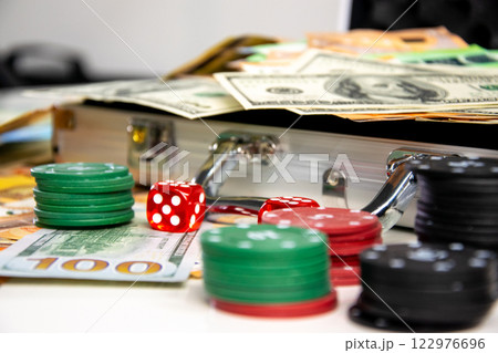 A close-up shot of a silver briefcase with red dice and stacks of casino chips and cash spilling out 122976696
