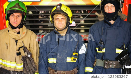 Portrait of male firefighters in helmets and protective uniforms standing near a big red car. Young confident firemen in full equipment posing against the background of a fire engine at station Portrait of male firefighters in helmets and protective uniforms standing near a big red car. Young confident firemen in full equipment posing against the background of a fire engine at station 122983267
