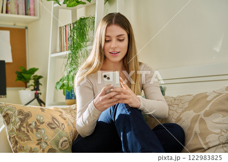Young smiling woman looking at smartphone sitting on couch at home Young smiling woman looking at smartphone sitting on couch at home 122983825