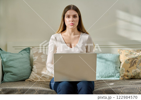 Beautiful young serious woman with laptop looking at camera sitting on couch at home Beautiful young serious woman with laptop looking at camera sitting on couch at home 122984570