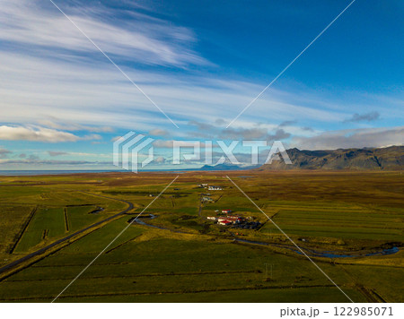 Breathtaking aerial perspective of the icelandic coastal countryside of the snaefellsnes peninsula, showcasing lush green fields, a meandering river, and distant mountains against a picturesque cloudy Breathtaking aerial perspective of the icelandic coastal countryside of the snaefellsnes peninsula, showcasing lush green fields, a meandering river, and distant mountains against a picturesque cloudy 122985071