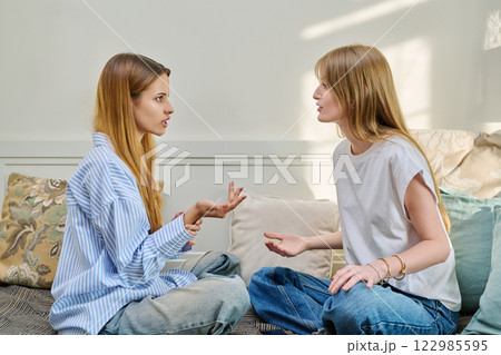 Two young serious teenage girls friends talking, sitting together on couch Two young serious teenage girls friends talking, sitting together on couch 122985595