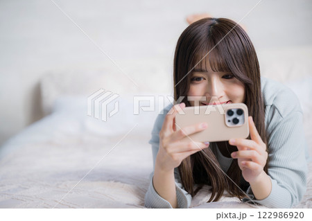 スマホを持って動画などを見るベッドで寝転ぶ笑顔の女性 スマホを持って動画などを見るベッドで寝転ぶ笑顔の女性 122986920