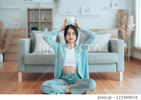 Young Asian woman holding piggy bank and sitting on the floor at home Young Asian woman holding piggy bank and sitting on the floor at home 122989018