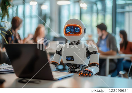 A robot is sitting in front of a laptop computer in a room with people 122990624