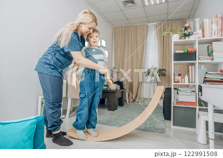 therapist assists boy balancing on wooden board in pediatric therapy room, Smiling, healthcare, education, pediatric services. Special needs education and support therapist assists boy balancing on wooden board in pediatric therapy room, Smiling, healthcare, education, pediatric services. Special needs education and support 122991508