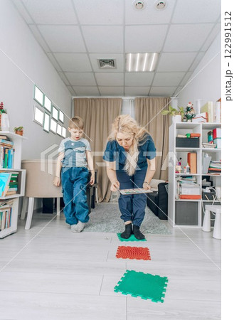 boy balancing on a red sensory mat while reaching for a card, session in bright room with therapist, concept of motor skill development, rehabilitation, pediatric physiotherapy boy balancing on a red sensory mat while reaching for a card, session in bright room with therapist, concept of motor skill development, rehabilitation, pediatric physiotherapy 122991512