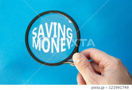Saving Money symbol. Concept words Saving Money on magnifying glass. Businessman hand. Beautiful blue background. Business and Saving Money concept. Copy space. 122991749