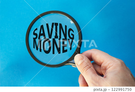 Saving Money symbol. Concept words Saving Money on magnifying glass. Businessman hand. Beautiful blue background. Business and Saving Money concept. Copy space. 122991750