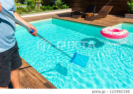 Man cleaning turquoise swimming pool with skimmer net, wooden deck, and colorful inflatable rings Man cleaning turquoise swimming pool with skimmer net, wooden deck, and colorful inflatable rings 122992198