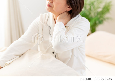 朝の寝室で首や肩のこりに苦しむ白いパジャマ姿のミドル女性 朝の寝室で首や肩のこりに苦しむ白いパジャマ姿のミドル女性 122992985