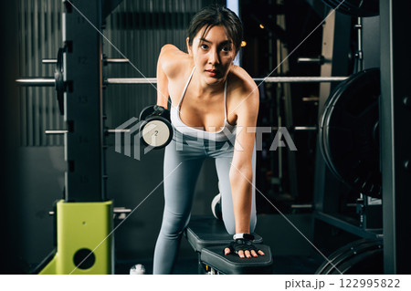 Asian woman lifting dumbbell on bench in fitness gym, Happy young female doing strength training exercise with dumbbells in a gym, Weight training healthy lifestyle concept Asian woman lifting dumbbell on bench in fitness gym, Happy young female doing strength training exercise with dumbbells in a gym, Weight training healthy lifestyle concept 122995822