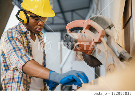 Construction worker in a yellow helmet using a chop saw to cut wood, with a vacuum cleaner collecting wood chips. woodworking, precision cutting, and construction sites, National Carpenters Day Construction worker in a yellow helmet using a chop saw to cut wood, with a vacuum cleaner collecting wood chips. woodworking, precision cutting, and construction sites, National Carpenters Day 122995833