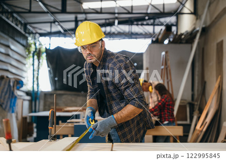 Carpenter wearing a yellow hard hat and safety glasses measuring wood with tape measure. Ideal for woodworking, furniture making, and cabinetry, showcasing precision and craftsmanship in the workshop 122995845