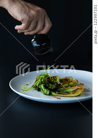 Hand sprinkles seasoning over freshly cooked bok choy on a sleek plate against a dark backdrop. Hand sprinkles seasoning over freshly cooked bok choy on a sleek plate against a dark backdrop. 122997101