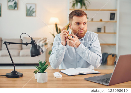 Holding the fruit. Man with banana sitting by the table with laptop on it and have fun Holding the fruit. Man with banana sitting by the table with laptop on it and have fun 122998619