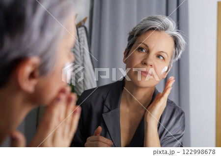 Mature beautiful woman during menopause with gray hair applies hormonal anti-aging cream to her face 122998678