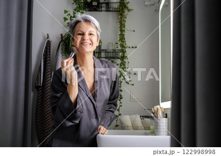 Mature happy aged beautiful woman during menopause with grey hair applying makeup on her face 122998998