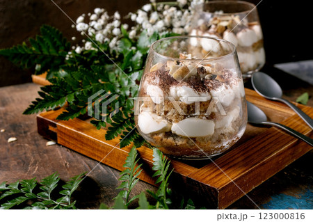 Classic Italian tiramisu layered dessert with cocoa powder and almonds in two glasses. Served on tray with spoons on old wooden table. Decorated white babys breath flowers and green leaves. Classic Italian tiramisu layered dessert with cocoa powder and almonds in two glasses. Served on tray with spoons on old wooden table. Decorated white babys breath flowers and green leaves. 123000816