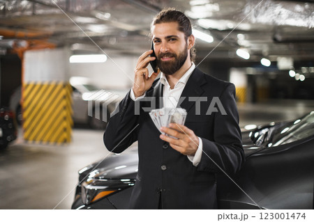 Young Caucasian male businessman in suit talking on phone holding cash in underground parking lot. 123001474
