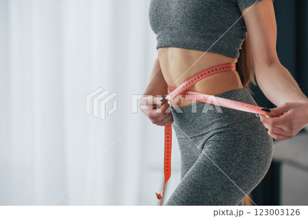 Pink colored measuring tape around the waist. Young woman with slim body type and in yoga clothes is at home 123003126